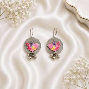 A pair of circular, oxidized silver hook earrings featuring a vibrant, hand-painted yellow bird motif with colorful wing accents, finished with small hanging silver ghungroo beads, displayed on white satin fabric.