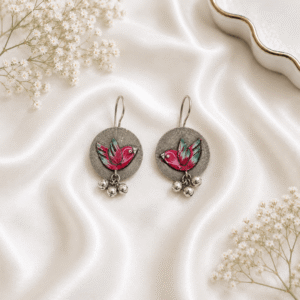 A pair of circular, oxidized silver hook earrings featuring a vibrant, hand-painted yellow bird motif with colorful wing accents, finished with small hanging silver ghungroo beads, displayed on white satin fabric.