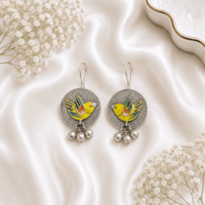 A pair of circular, oxidized silver hook earrings featuring a vibrant, hand-painted yellow bird motif with colorful wing accents, finished with small hanging silver ghungroo beads, displayed on white satin fabric.