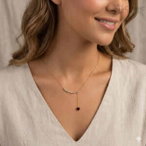 A delicate gold-toned lariat necklace worn by a woman, featuring a curved arc of sparkling clear crystals and a vertical drop chain ending in a small, deep red heart-shaped pendant.