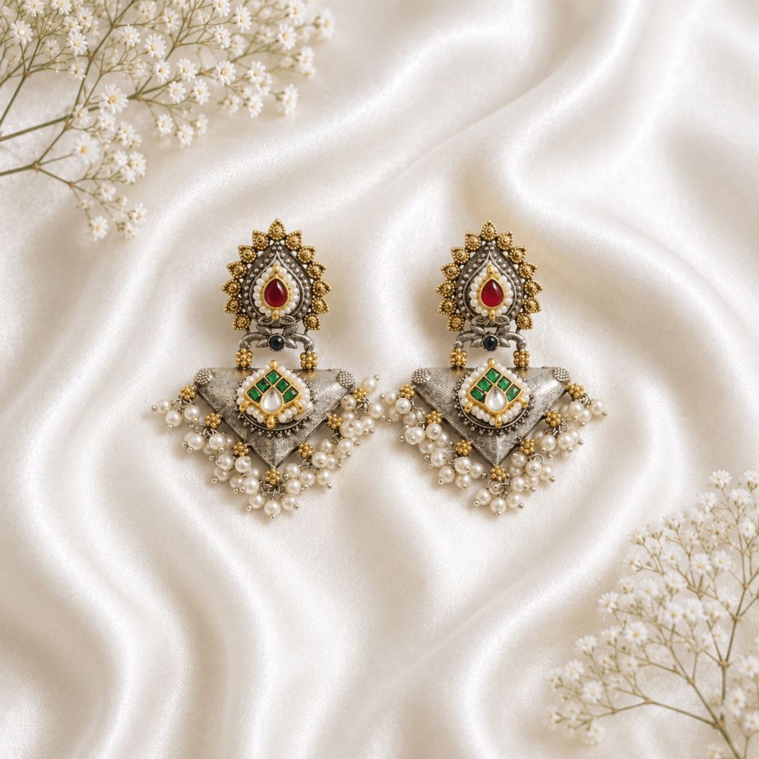 Pair of Kreezo antique dual-tone statement earrings featuring intricate gold filigree, red and green gemstone accents, Kundan work, and white pearl clusters on a clean white background.