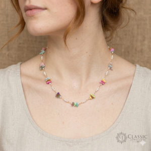 A close-up view of a delicate gold-chain necklace worn by a woman with red hair, featuring small white pearls interspersed with varied multi-colored polished stone chips (pink, yellow, green, teal, purple).