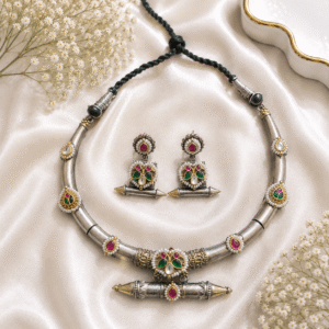 An antique silver-toned Hasli necklace and matching drop earrings set featuring multicolor Kundan, ruby red, and emerald green glass stones with delicate pearl borders, displayed on a cream satin background.