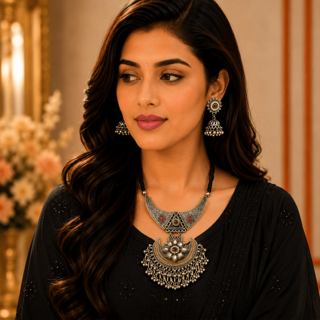 A woman wearing an intricate oxidized silver statement necklace with a crescent pendant and matching dangling jhumka earrings, adorned with red and green stones.