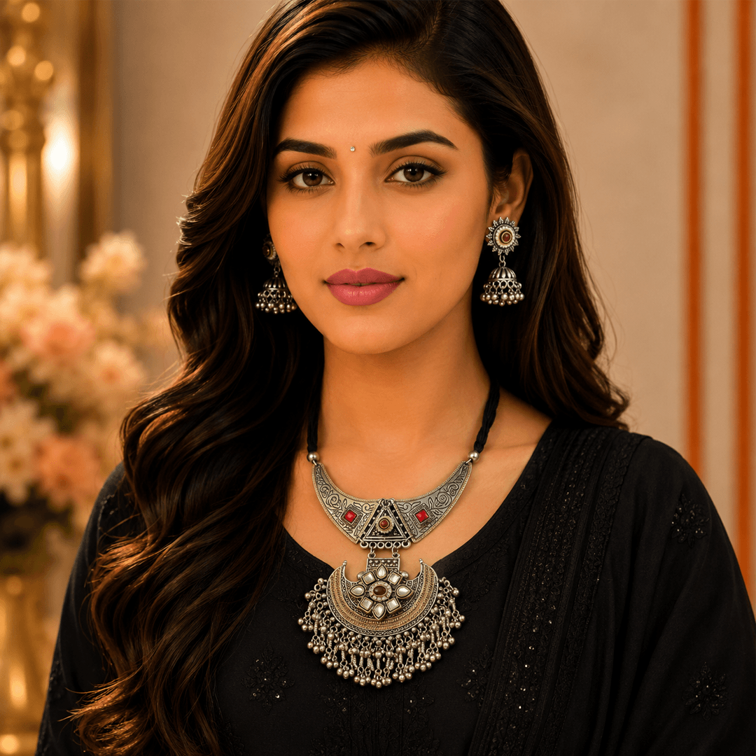 A woman wearing an intricate oxidized silver statement necklace with a crescent pendant and matching dangling jhumka earrings, adorned with red and green stones.