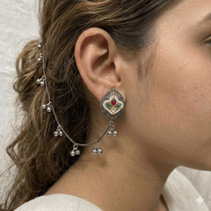 Close-up of a woman wearing an antique oxidized silver stud earring featuring red and green kemp stones, connected to a long hair chain or kaan chain adorned with tiny hanging silver ghungroo bells.