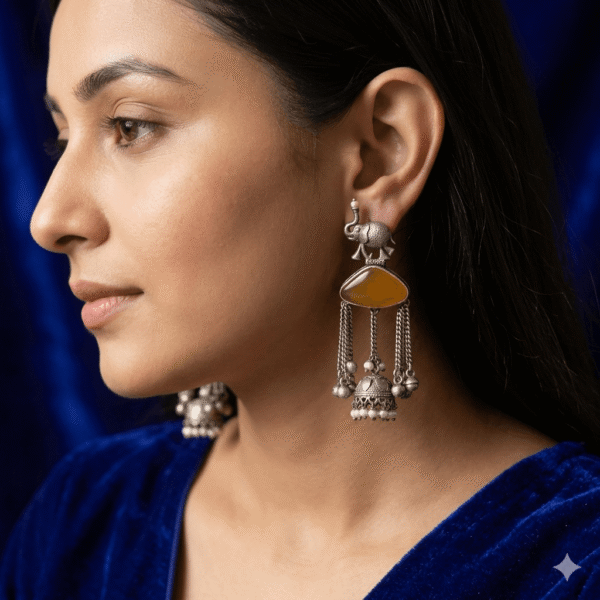 Model wear A pair of handcrafted German silver statement earrings featuring a detailed elephant motif at the top, a triangular amber-colored gemstone center, and hanging chain tassels with tiny silver bells and a small jhumka with pearl beads