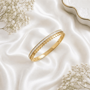 A refined gold-tone hinged kada bracelet featuring a row of sparkling baguette-cut stones bordered by a delicate, repetitive scalloped design, resting on a soft white silk fabric background.