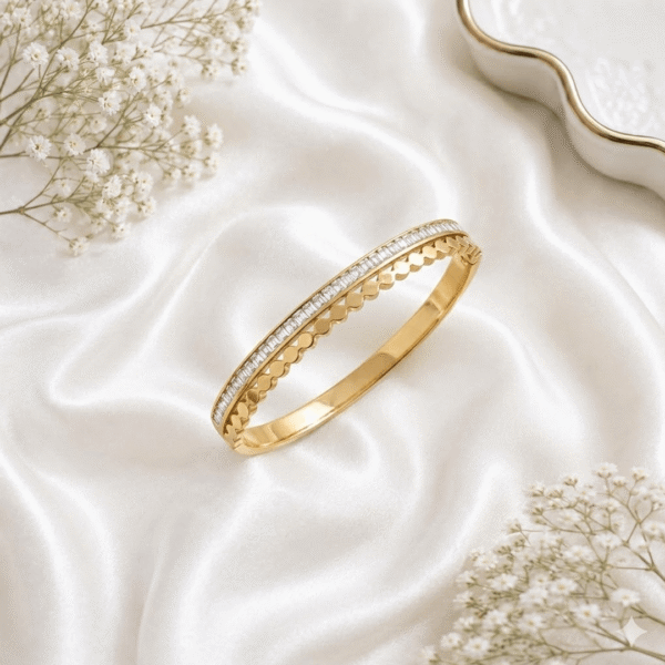 A refined gold-tone hinged kada bracelet featuring a row of sparkling baguette-cut stones bordered by a delicate, repetitive scalloped design, resting on a soft white silk fabric background.