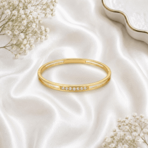 A sleek, gold-plated open-hinge kada bracelet featuring a minimalist design with a single row of five small shimmering crystals, resting on white satin fabric.
