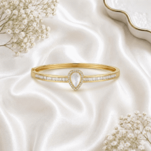 A luxurious gold-tone bangle bracelet featuring a central teardrop-shaped mother-of-pearl stone surrounded by a halo of micro-stones, flanked by a row of baguette-cut stones, set against a soft white satin background.