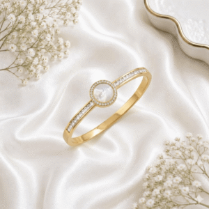 An elegant gold-plated open-hinge kada bracelet featuring a central round Mother of Pearl stone encircled by small crystals, flanked by rows of baguette-cut crystals on the band, resting on white satin fabric.