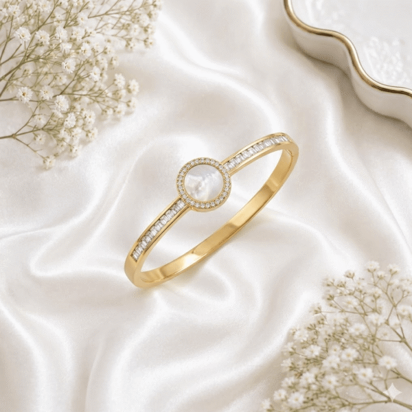 An elegant gold-plated open-hinge kada bracelet featuring a central round Mother of Pearl stone encircled by small crystals, flanked by rows of baguette-cut crystals on the band, resting on white satin fabric.