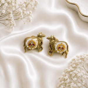 Pair of antique brass-finish Kamadhenu cow earrings adorned with white pearls and red ruby stones, displayed on a white satin fabric background with white flowers.