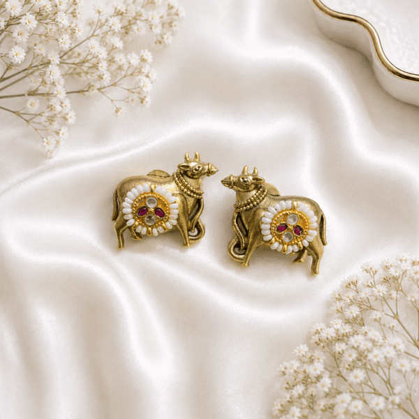 Pair of antique brass-finish Kamadhenu cow earrings adorned with white pearls and red ruby stones, displayed on a white satin fabric background with white flowers.