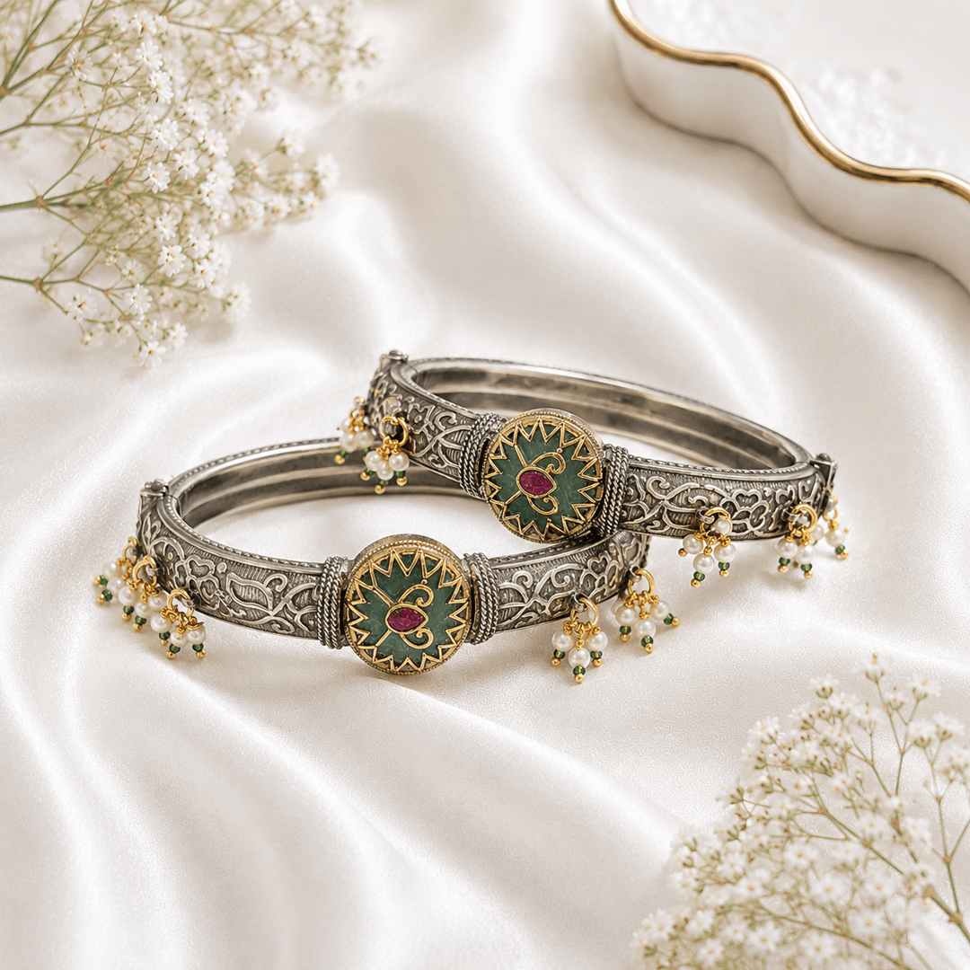 A pair of ornate dual-tone heritage bangles featuring intricate silver engraving, a central green and gold enamel medallion with a ruby center, and delicate pearl drop charms, displayed on a soft white satin background.