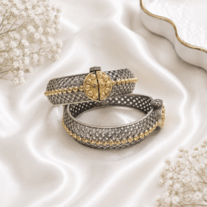 A pair of intricate dual-tone, silver and gold-plated filigree cuff bangles displayed on a soft, white satin background with delicate white flowers.