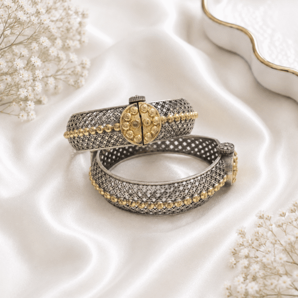A pair of intricate dual-tone, silver and gold-plated filigree cuff bangles displayed on a soft, white satin background with delicate white flowers.