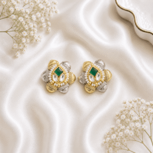 A pair of ornate dual-tone (gold and silver) floral-shaped stud earrings featuring a central square green stone, surrounding teardrop-shaped white stones, and an inner border of small white pearls, resting on white satin.