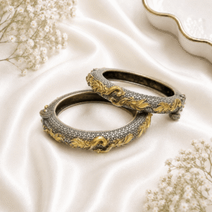 A pair of ornate dual-tone hinged bangles featuring a textured silver base with intricate, flowing golden peacock motifs, displayed on a soft white satin background.