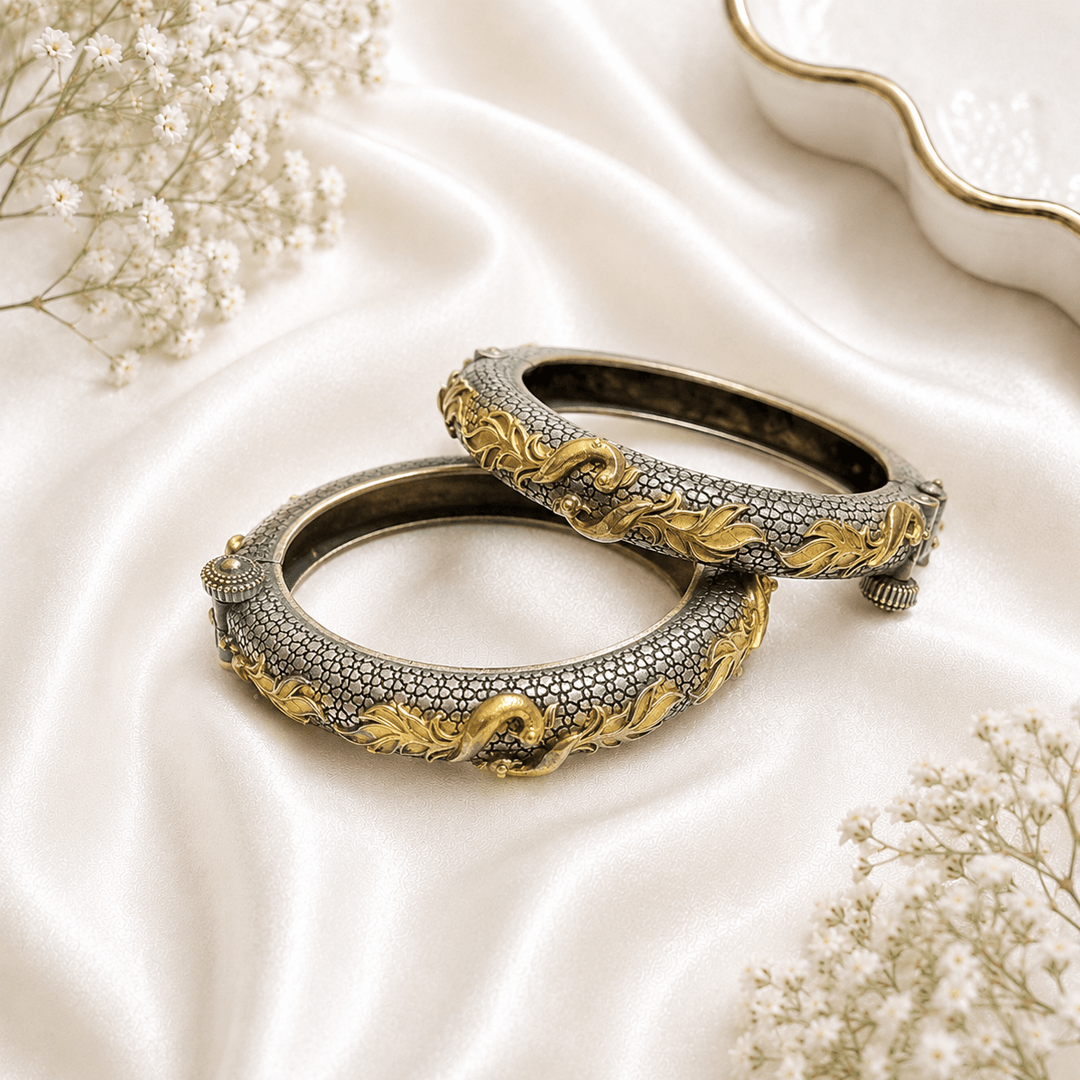 A pair of ornate dual-tone hinged bangles featuring a textured silver base with intricate, flowing golden peacock motifs, displayed on a soft white satin background.