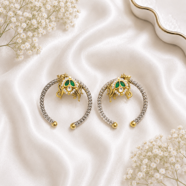 A pair of traditional hoop earrings crafted in German silver with gold-tone accents, featuring a Nandi (bull) motif adorned with a green teardrop stone and pearl detailing, resting on white satin.