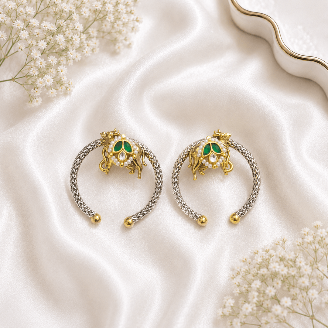 A pair of traditional hoop earrings crafted in German silver with gold-tone accents, featuring a Nandi (bull) motif adorned with a green teardrop stone and pearl detailing, resting on white satin.