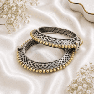 A pair of hinged bangles featuring a silver-tone cross-hatch lattice design framed by rich golden beaded borders, elegantly placed on white satin.