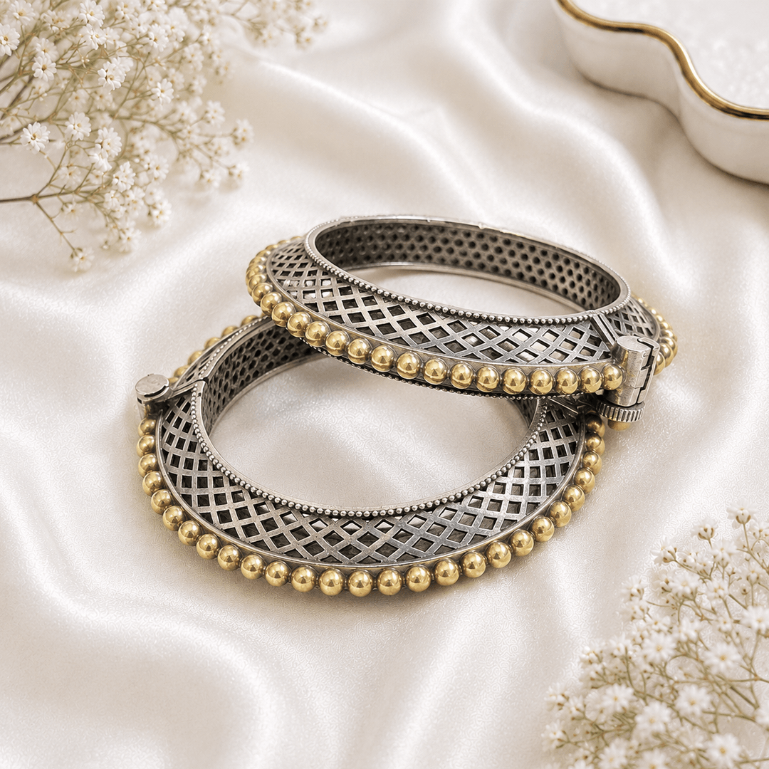 A pair of hinged bangles featuring a silver-tone cross-hatch lattice design framed by rich golden beaded borders, elegantly placed on white satin.