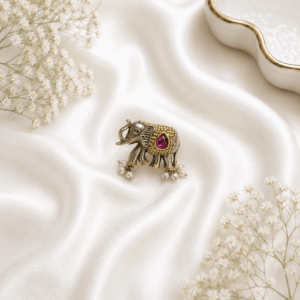 A detailed view of a uniquely designed, german silver and gold-tone ring shaped like an elephant, featuring a prominent pink teardrop gemstone and small delicate pearl dangles, resting on a soft white silk fabric background.