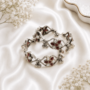 A pair of intricate, floral-inspired german silver bangles featuring ruby-red marquise stones, white pearls, and dangling metallic ghungroo bells, arranged on a white satin background.