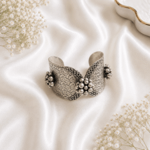 A wide, open-ended German silver cuff kada featuring intricate etched floral patterns and clustered metallic bead details, resting on a draped white satin background.