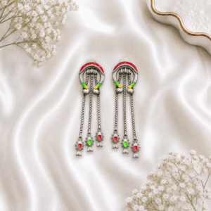 A pair of long, artistic silver dangle earrings featuring a curved upper arch with colorful enamel details and three hanging chains ending in tiny fish-shaped charms with red and green enamel, resting on white satin fabric.