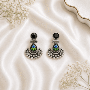 A pair of German silver earrings featuring a round deep red stone top and a teardrop-shaped base decorated with a delicate black floral motif, finished with granulated metal detailing and set against a white satin background.