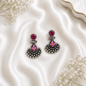 A pair of German silver earrings featuring a round deep red stone top and a teardrop-shaped base decorated with a delicate pink and yellow floral motif, finished with granulated metal detailing and set against a white satin background.