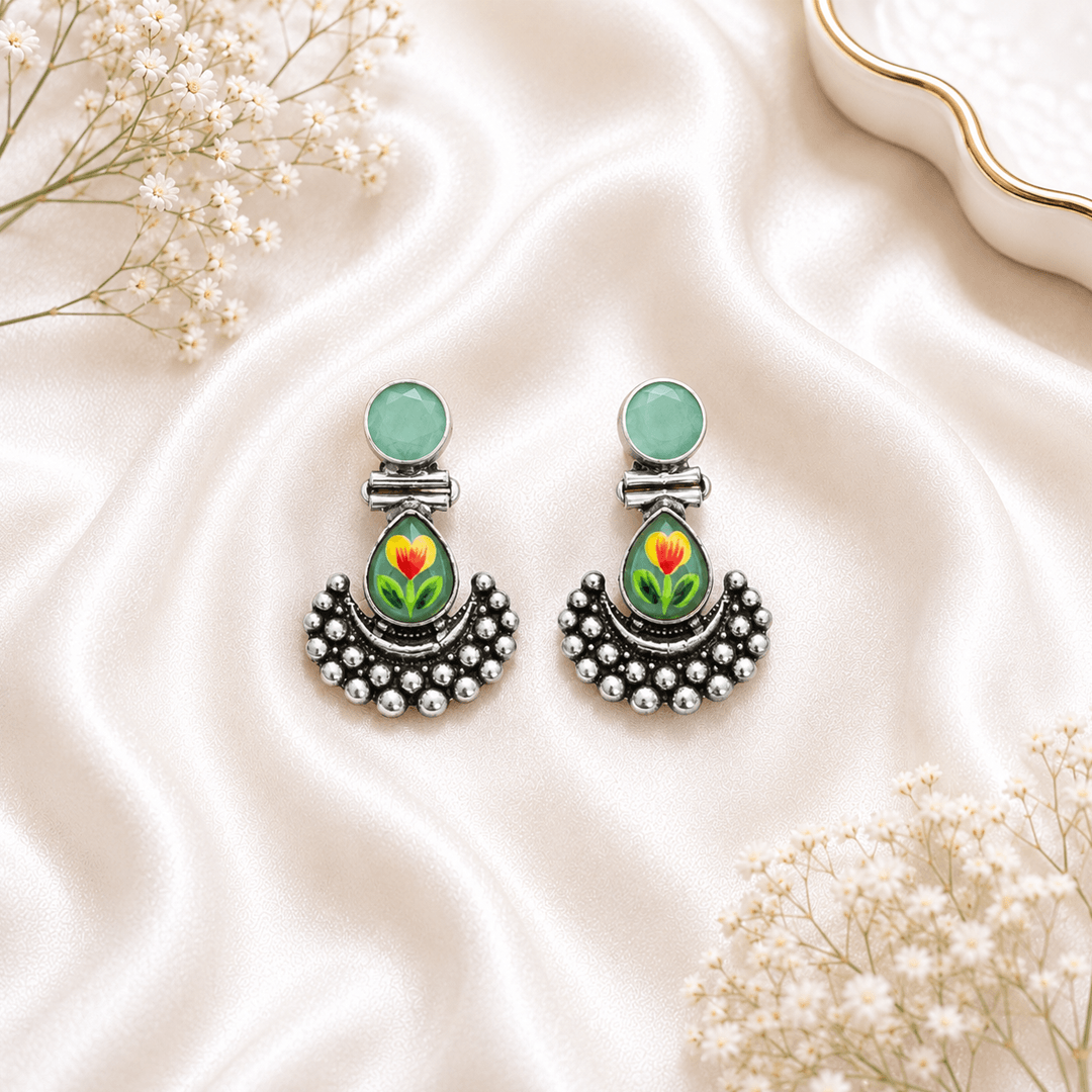 A pair of German silver earrings featuring a round deep red stone top and a teardrop-shaped base decorated with a delicate green floral motif, finished with granulated metal detailing and set against a white satin background.