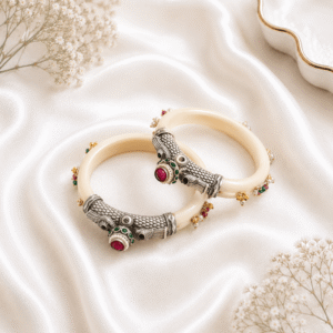 A pair of elegant kadas featuring an ivory-look base, intricately designed German silver caps, and vibrant red and green stone embellishments, resting on a soft white silk background.