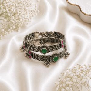 A pair of hinged silver-tone bangles featuring a complex braided metal weave, adorned with central green stone stations and pink accents, finished with delicate dangling ghungroo bells, displayed on a white satin background.