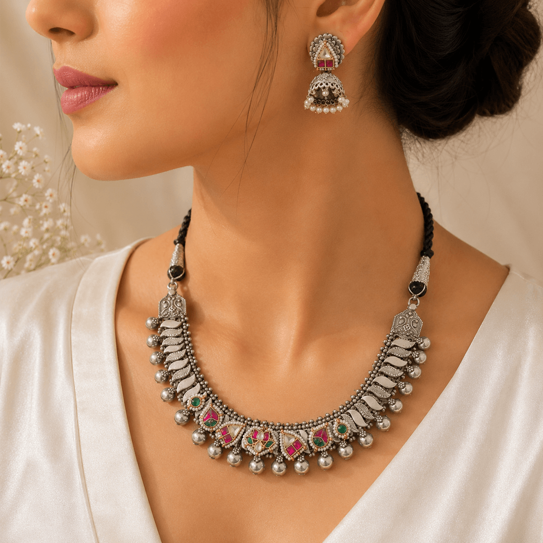A premium German silver and Panchi Kundan jewelry set featuring a necklace with intricate leaf-like designs and multicolored stone accents, accompanied by matching jhumka earrings, displayed on a textured white fabric with floral elements.