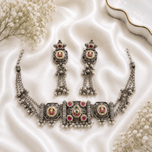 A detailed view of an exquisite German silver necklace and matching earring set featuring traditional Panchi Kundan work, vibrant red ruby-toned stones, and delicate pearl drop embellishments, displayed on white satin fabric.