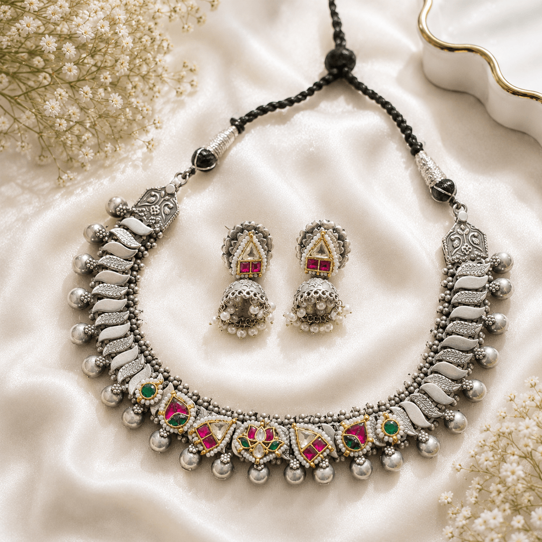 A premium German silver and Panchi Kundan jewelry set featuring a necklace with intricate leaf-like designs and multicolored stone accents, accompanied by matching jhumka earrings, displayed on a textured white fabric with floral elements.