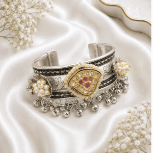 An ornate, wide German silver cuff kada featuring an intricate central medallion with pink stones, flanked by pearl clusters and finished with dangling metallic ghungroo bells, displayed on white satin.