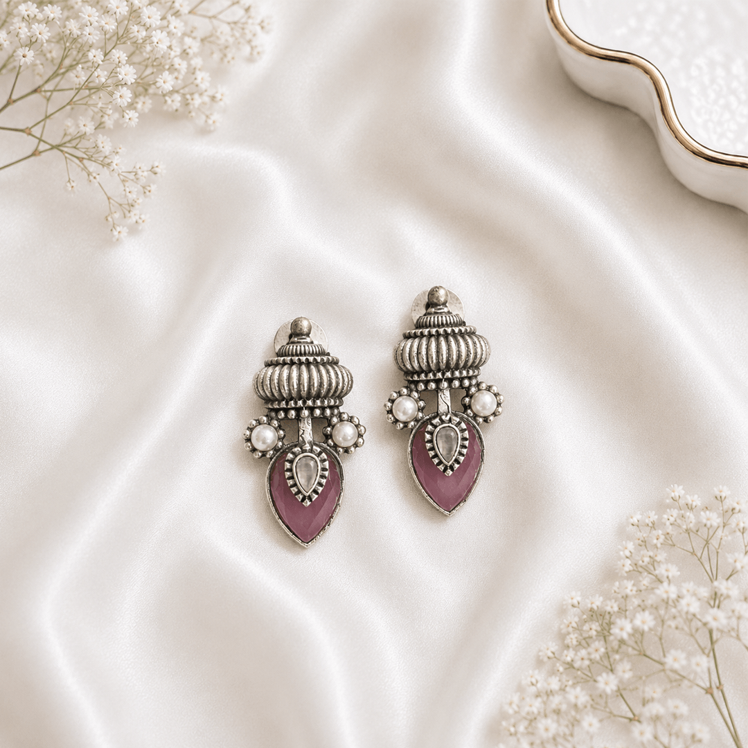 A pair of handcrafted German silver drop earrings featuring a decorative dome top, accented with white pearls, and a teardrop-shaped pink gemstone center, resting on soft white silk fabric.