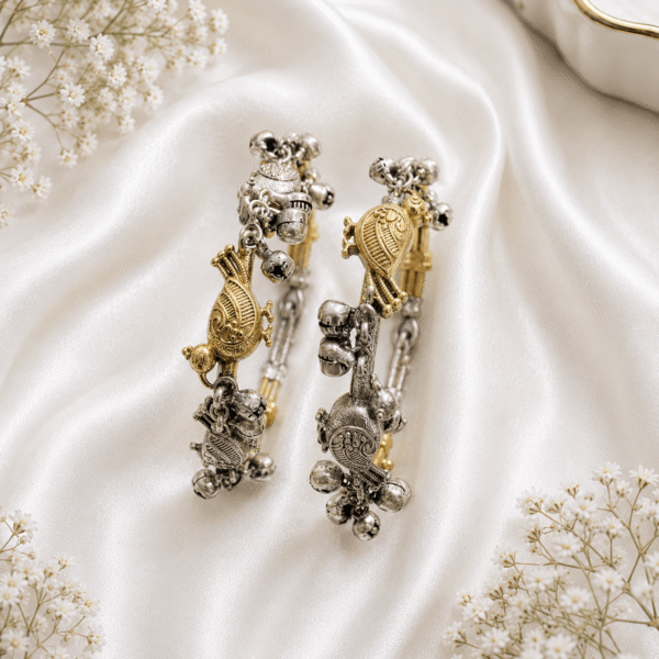 A pair of dual-tone silver and gold-plated Kada style bangles featuring intricate bird and tortoise charms with dangling silver ghungroo bells, displayed on a draped white satin background.