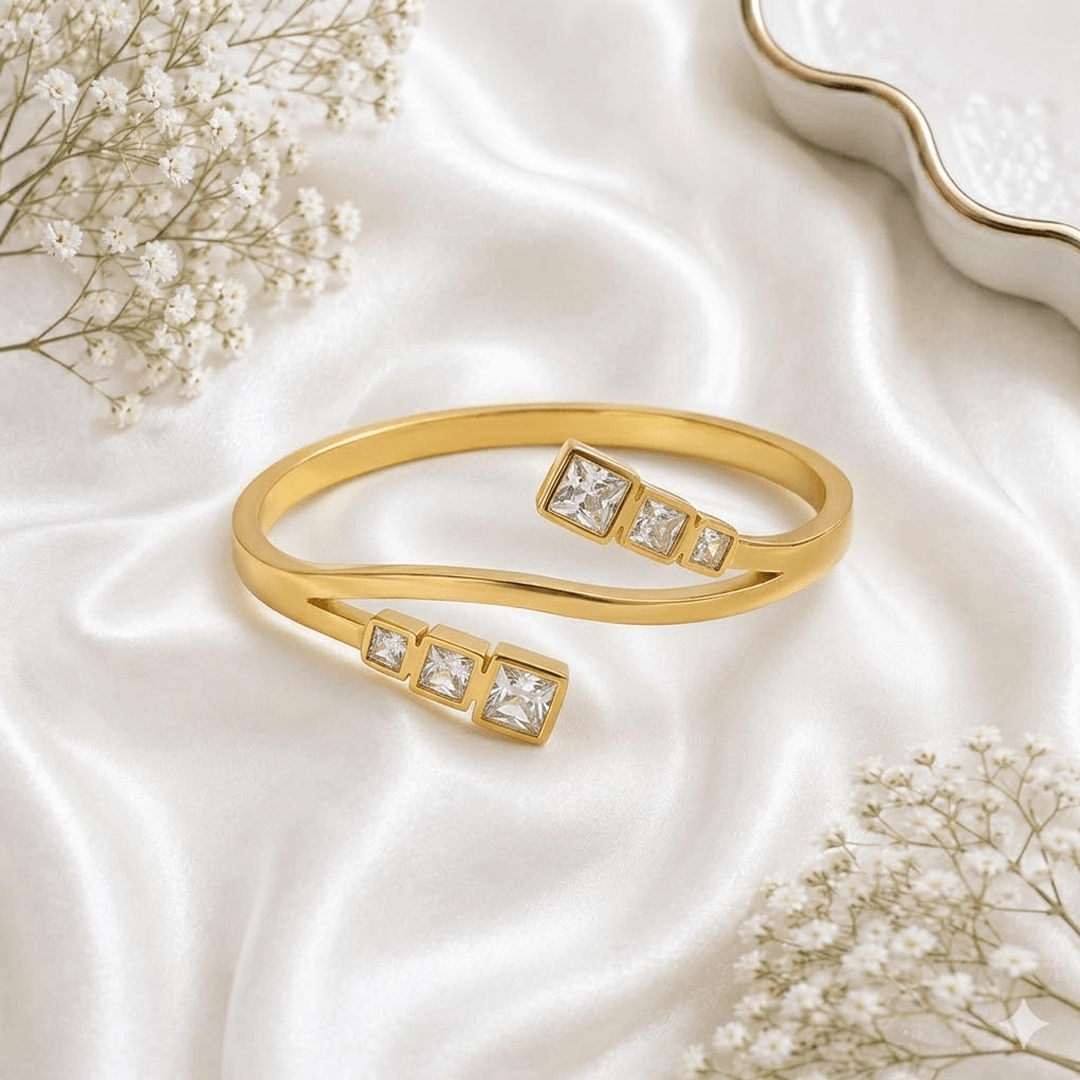 A sleek, open-style gold-tone kada bracelet featuring three small square-cut sparkling zircon stones at each terminal, resting elegantly on soft white silk fabric.