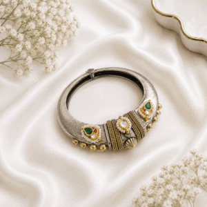 A single, wide, german silver-tone kada bracelet featuring an ornate central section with Kundan stone settings, green and red accents, pearls, and dangling metallic beads, resting on a white satin background.