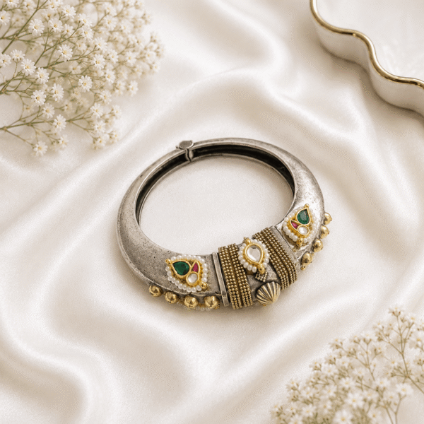 A single, wide, german silver-tone kada bracelet featuring an ornate central section with Kundan stone settings, green and red accents, pearls, and dangling metallic beads, resting on a white satin background.