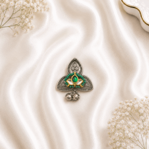A detailed top-down view of an intricate, oxidized silver-tone ring featuring a stylized lotus design with a central green gemstone, surrounded by small green beads and finished with dangling metallic ghungroo charms, resting on a draped white silk background.