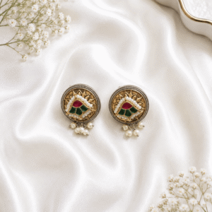 Elegant woman wearing round gold-plated traditional Indian stud earrings featuring red and green Meenakari work and hanging pearl drops.