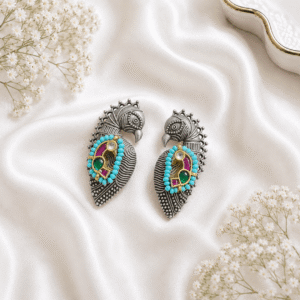 A pair of German silver peacock-shaped statement earrings featuring turquoise beads, green, and magenta pink stones, displayed on a soft white silk fabric background.